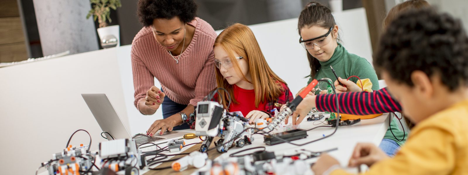 Group of happy kids with their African American female science teacher with laptop programming electric toys and robots at robotics classroom